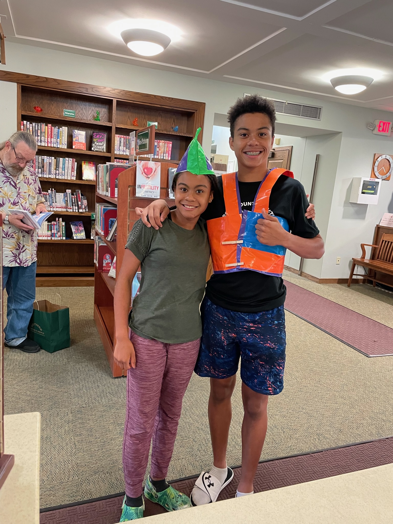A boy and a girl stand wearing a duct tape hat and vest.